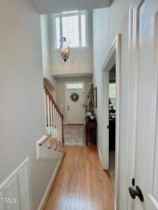 A hallway with light wood floors leads to a white front door with a wreath. A staircase with white spindles is on the left, and a chandelier hangs from a high ceiling above the entryway. A room is visible on the right.