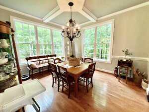 A dining room with hardwood floors, a wooden table and six chairs, a bench by large windows, a chandelier, and various decorative items and furniture along the walls. Trees are visible through the windows.