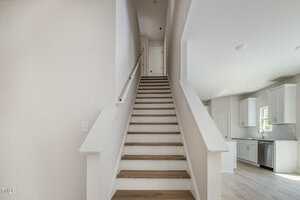 A straight staircase with light wood steps and white railings leads up to a closed door. The adjacent kitchen features white cabinets, a window, and light flooring. The walls are painted a light, neutral color.