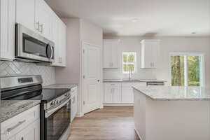 A modern kitchen with white cabinets, stainless steel appliances, a light gray herringbone tile backsplash, granite countertops, light wood flooring, and large windows letting in natural light.