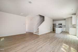 Open-concept living space with light wood flooring, white walls, and a staircase leading upstairs. The kitchen features white cabinets, stainless steel appliances, and an island. Natural light enters through several windows.