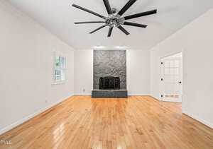 A spacious, empty living room with light wood floors, white walls, a large black ceiling fan, a stone fireplace, and doors with glass panels. One window with white shutters is on the left wall.
