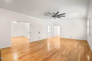 Spacious empty room with light wood flooring, white walls, large black ceiling fan, and multiple doorways leading to other rooms. Natural light comes through windows with white shutters.