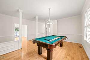 A room with light wood floors and white walls features a pool table centered under a modern chandelier. Natural light enters through large windows with white shutters.