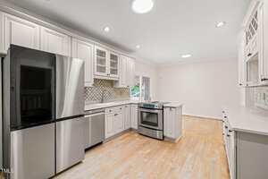 A modern kitchen with white cabinets, stainless steel appliances, light wood flooring, a small center island, and a tile backsplash. Recessed lighting and a round ceiling light illuminate the space.