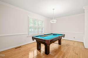 A room with light-colored walls and hardwood floors features a pool table with a green felt top centered under a modern pendant light, next to a window with white plantation shutters.
