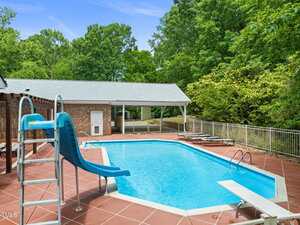 Outdoor swimming pool with a blue slide, diving board, and lounge chairs on a red-tiled deck at 1514 Lawrence Road in Creedmoor. A metal fence and trees surround the area, with a brick building and covered patio visible in the background.