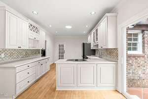 A bright kitchen with white cabinets, light countertops, a center island with a cooktop, tile backsplash, stainless steel appliances, wood flooring, and a glass door leading outside.