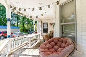 A covered wooden porch with string lights, a round cushioned chair, a rustic table, and a large window. Several cars are parked outside next to trees and a blue-roofed building.