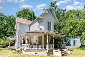 A two-story white wooden house with a covered wraparound porch, decorative trim, and surrounded by green trees, photographed on a sunny day with blue sky and scattered clouds.