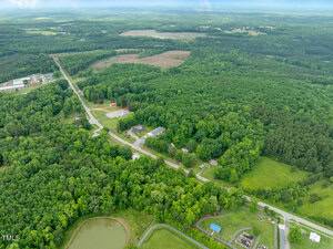 Aerial view of a rural landscape with dense green forest, scattered houses, a pond, open fields, and intersecting roads stretching through the area under a cloudy sky.