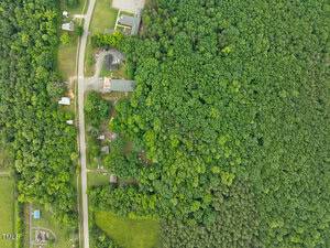 Aerial view of a forested area with a road running vertically along the left side, residential buildings, and cleared green spaces; dense trees cover most of the right side of the image.