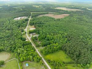 Aerial view of a rural area with dense green forest, a winding road, scattered houses, a pond, and open fields. Buildings are visible in the distance, surrounded by lush vegetation and trees.
