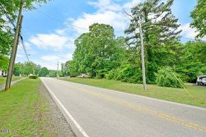 A two-lane road runs through a suburban area with grass shoulders, utility poles, and trees on both sides. Houses and vehicles are visible in the background under a partly cloudy sky.