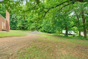 A dirt and gravel driveway curves beside a red brick building on the left, with a grassy area and large trees overhead. In the background, there are piles of gravel and a white trailer near dense green woods.