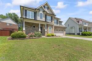 A two-story suburban house with a brick and siding exterior, a covered front porch, attached two-car garage, and a lawn with landscaped flower beds. Other homes and a clear sky are visible in the background.