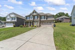 A two-story suburban house with brick and siding exterior, three-car garage, and a wide concrete driveway. The lawn is green but patchy, and neighboring houses are visible on both sides. The sky is partly cloudy.