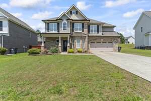 A two-story suburban house with a brick and siding exterior, attached two-car garage, front porch with columns, and a well-kept lawn on a sunny day. Neighboring houses are visible on either side.