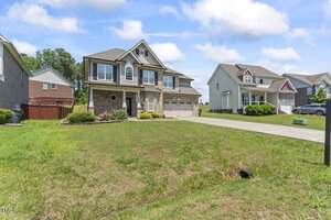 A two-story suburban house with a stone and siding exterior, double garage, and a well-maintained front yard. Neighboring homes and a concrete driveway are visible under a partly cloudy sky.