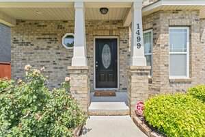 A brick house exterior with a black front door featuring a glass oval panel, white columns, house number 1499, a small window, and green bushes with flowers beside the entrance walkway.