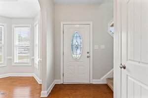 A white front door with an oval glass panel is centered in an entryway with light-colored walls and wooden floors. To the left are three windows, and to the right are stairs carpeted in beige.