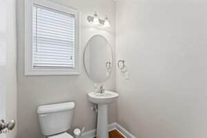 A small bathroom with a white toilet, a pedestal sink, an oval mirror, a wall-mounted light fixture, and a window with white blinds. The walls are light beige, and the floor has a wood-like finish.