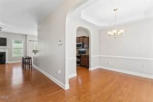 An open interior view of a home showing a dining area with a chandelier, hardwood floors, and arched entryway leading to a kitchen with dark cabinets and stainless steel appliances. A living area is visible in the background.