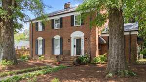 Nestled at 925 S Chestnut Street in Henderson, this two-story brick house features black shutters, a white arched doorway, and a charming porch. The yard boasts large trees and landscaped paths with mulch and plants.