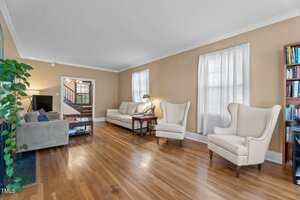A spacious living room with hardwood floors, beige walls, two large windows with white curtains, two armchairs, two sofas, a coffee table, a bookcase, a houseplant, and a TV. A stairway is visible in the background.
