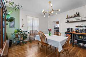A dining room with a rectangular table covered by a white tablecloth, wooden chairs, a modern chandelier, potted plants in the corner, and a shelving unit with coffee makers and various items on the right wall.