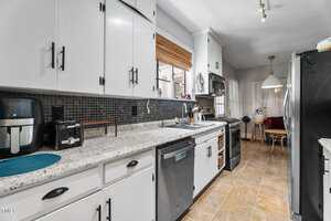 A modern kitchen with white cabinets, black hardware, granite countertops, stainless steel appliances, tiled floor, and a small dining area with a round table and chairs in the background.