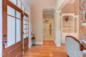 A home entryway with a wooden front door and glass panels, hardwood floors, beige walls, a small table with a lamp, built-in shelves, and a partial view of a stone fireplace and sofa.