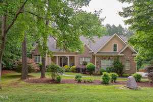 A two-story suburban house with tan siding and brick accents sits at 504 Belmont Circle, Wake Forest. Surrounded by trees and landscaped shrubs, the home features a grassy yard with a stone near the front and a partially visible driveway on the right.