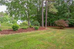 A landscaped yard with green grass, mulch, shrubs, and several mature trees. In the background, a white house is partially visible among trees, and the area appears well-maintained and bordered by woods.