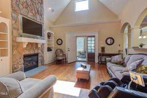A living room with a stone fireplace and mounted TV, built-in shelves, wood floors, large windows, a sofa, armchair, and massage chair. French doors lead to a sunroom; the kitchen is visible through an arched opening.