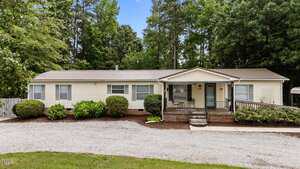 Charming single-story beige manufactured home at 2620 Hicksboro Road, Henderson, featuring a metal roof, front porch with ramp and steps, surrounded by shrubs and trees, and a gravel driveway in front.