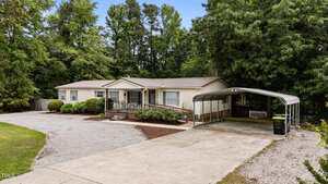 Single-story beige house with a metal roof, front porch with railing, gravel driveway, and attached carport on the right. The yard is bordered by trees and bushes, with a wooded area in the background.