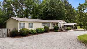 Single-story beige manufactured home with a metal roof, front porch, and ramp. Gravel driveway leads to a carport. The yard has bushes, trees, and a wooden fence to the left. Dense trees are in the background.