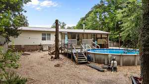 A beige manufactured home with a covered porch is shown in a wooded backyard. An above-ground pool with a deck and stairs is in the center, and the ground is covered with gravel. Trees surround the area.