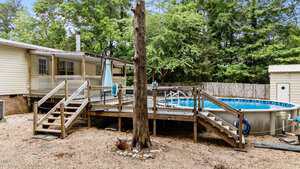 Backyard with a raised wooden deck featuring two staircases, an above-ground pool with railing, a tree in front, gravel ground cover, and a fenced yard with trees and a shed in the background.