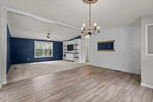 A living area with a tiled floor transitions into a carpeted section. The room has blue and white walls, a ceiling fan, a chandelier, large window, built-in shelving, and a wooden-framed niche on the wall.