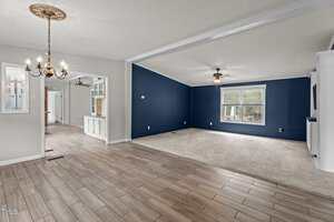 Open living area with tile and carpet flooring, navy accent wall, ceiling fans, white trim, and a chandelier. A dining area leads to a bright hallway and kitchen with glass-front cabinets in the background.