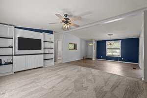 A spacious living room with built-in white shelves and a TV on the left, beige carpet, a ceiling fan, and an adjoining dining area with blue accent walls, a window, and tile flooring.