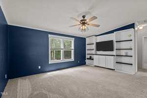 A room with blue accent walls, beige carpet, a ceiling fan with a light, a large window, and built-in white shelving and cabinets with a mounted TV. The room is empty of furniture.