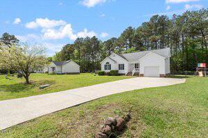 A white single-story house with a two-car garage and large driveway sits on a grassy lot with trees in the background. Another similar house is visible nearby under a blue sky with scattered clouds.