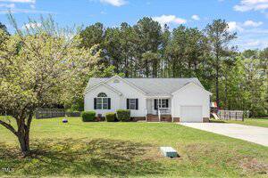 A single-story white house with a front porch, attached ramp, and manicured bushes sits on a grassy lawn with a tree and woods in the background. A concrete driveway leads up to the house.