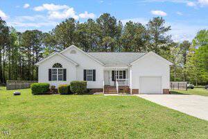 A single-story white house with black shutters, a front porch with steps, an attached single-car garage, manicured bushes, and a large grassy lawn, with trees and a wooden fence in the background.