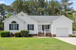 A single-story white house with black shutters, a front porch with steps, and a one-car garage sits on a well-kept lawn at 1752 Cobblestone Drive in Creedmoor, framed by tall pine trees.