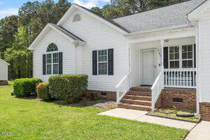 A single-story white house with black shutters, a brick foundation, and a small covered front porch with white railings. There are trimmed bushes and a lawn in front, with trees in the background.