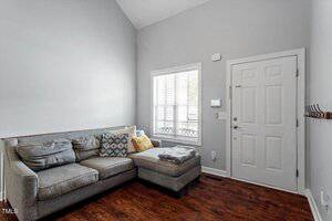 A small living room with a gray sectional sofa, assorted pillows, and a blanket. There is a white front door, a window with blinds, a wall-mounted coat rack, and wood flooring. The walls are light gray.